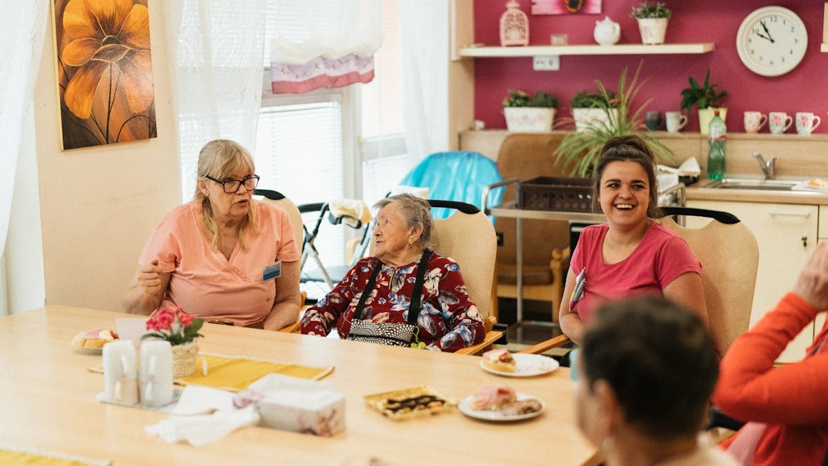 Elderly residents and caregivers enjoying a meal and conversation in a cheerful dining space.